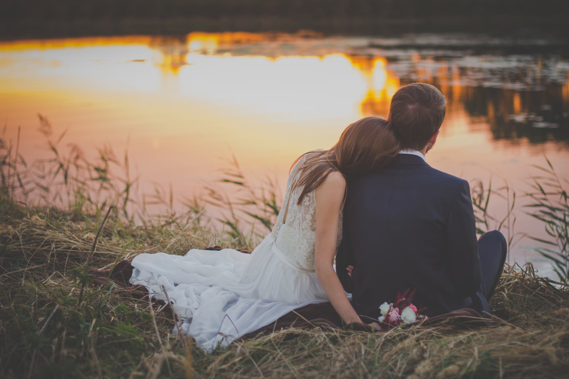 wedding couple sitting on green grass in front of body of water at sunset