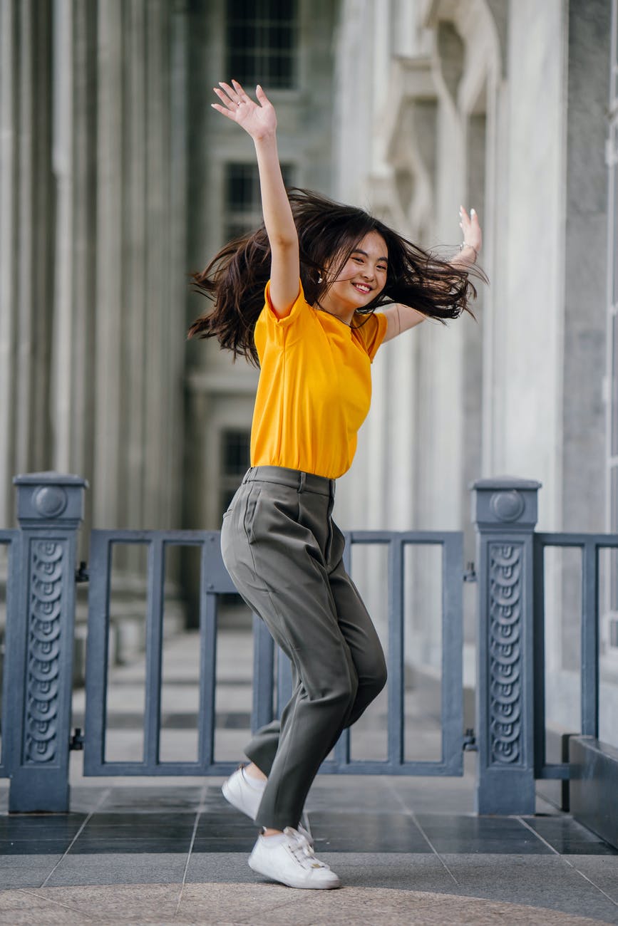 photo of smiling woman in yellow t shirt and grey pants with her hands raised